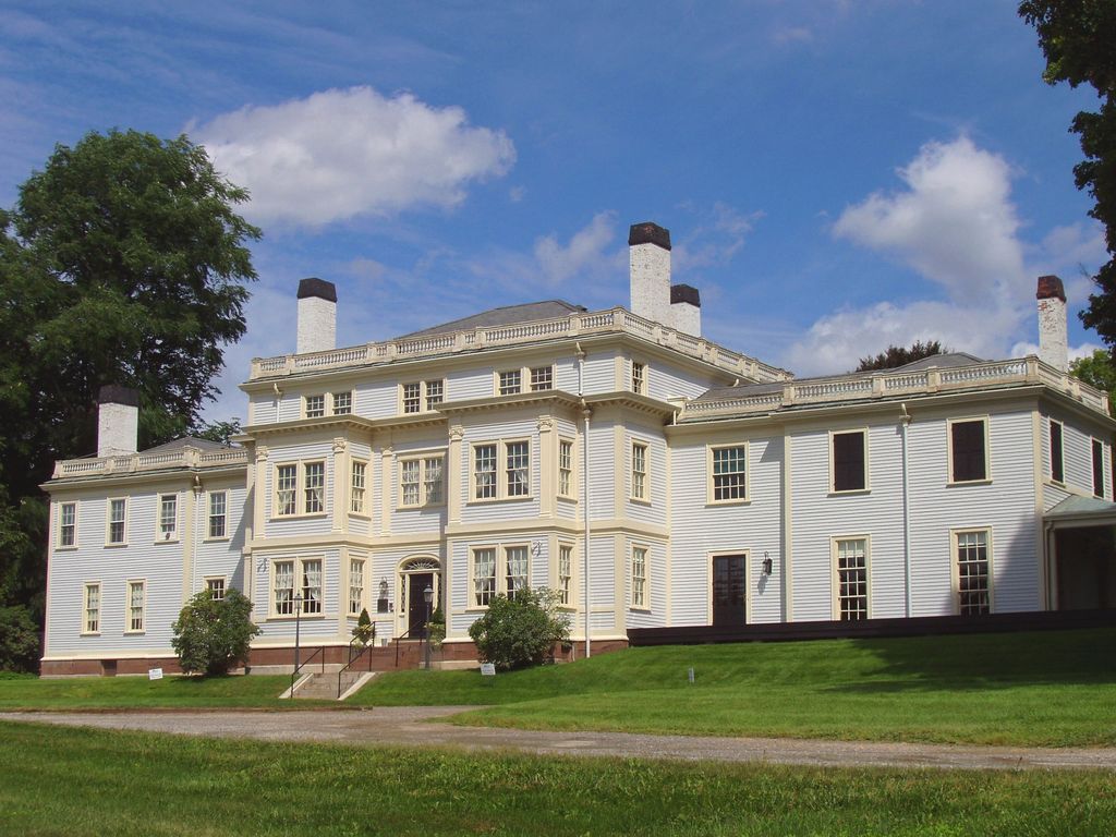 A long pale clapboard house has a three-storey centre, projecting window bays, lower side ranges, chimneys and roof balustrades.