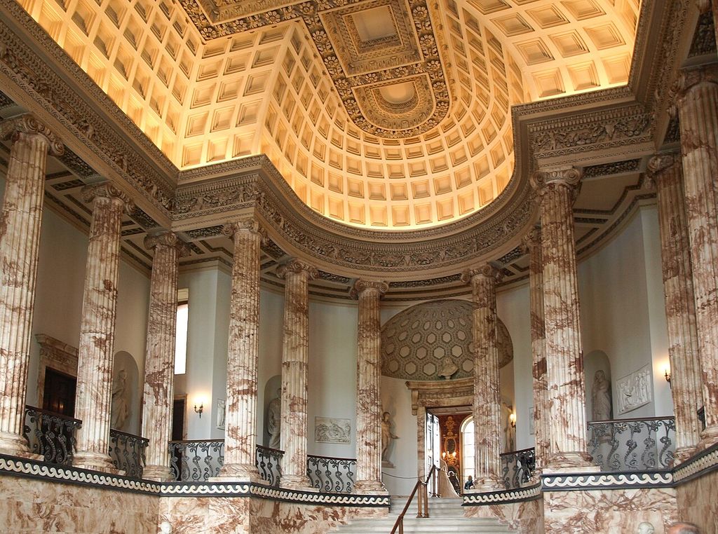 A broad stair rises between tall veined columns beneath an illuminated coffered ceiling and richly carved entablature.