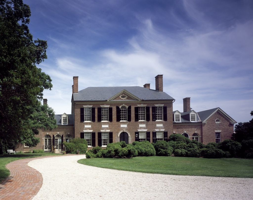 A brick country house with a central arched entrance is joined to lower wings beyond a curved drive and dense shrubs.