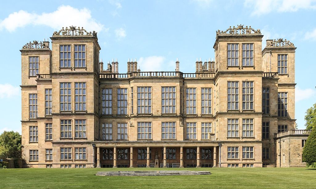 A symmetrical stone country house has projecting towers, dense grids of tall windows and ES initials along its parapets.