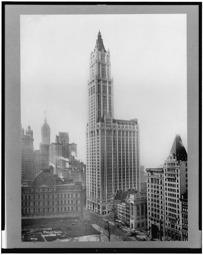 A historical black-and-white city view shows a very tall Gothic-crowned tower above lower buildings, streets and a large excavated site.