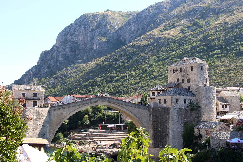 A single pale stone arch with a steep deck spans a rocky river between masonry towers, town buildings and steep mountains.