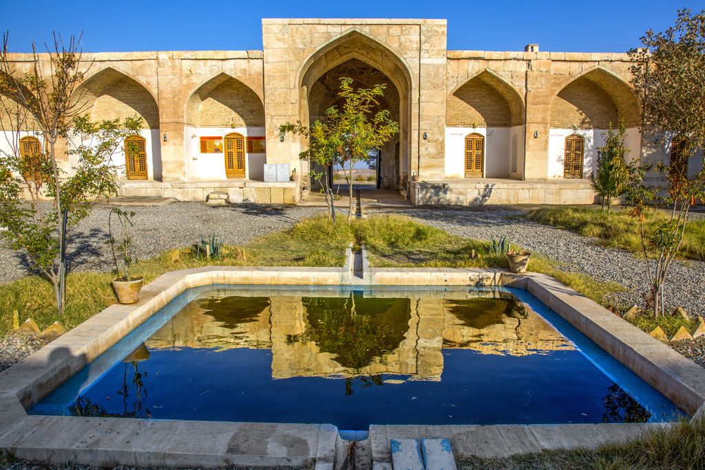 A pool and small planted beds face a central pointed entrance and repeated arched recesses containing doors around a caravanserai courtyard.