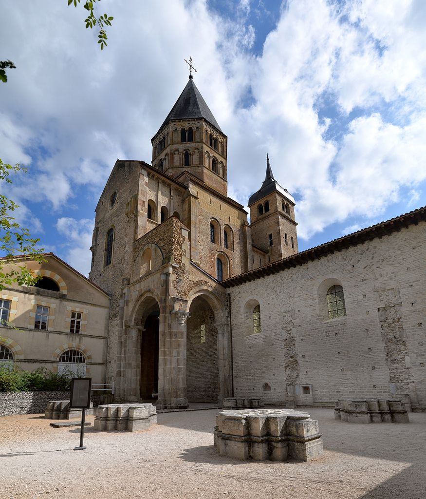 A tall Romanesque transept fragment and octagonal tower rise above low exposed stone bases in a gravelled archaeological area.