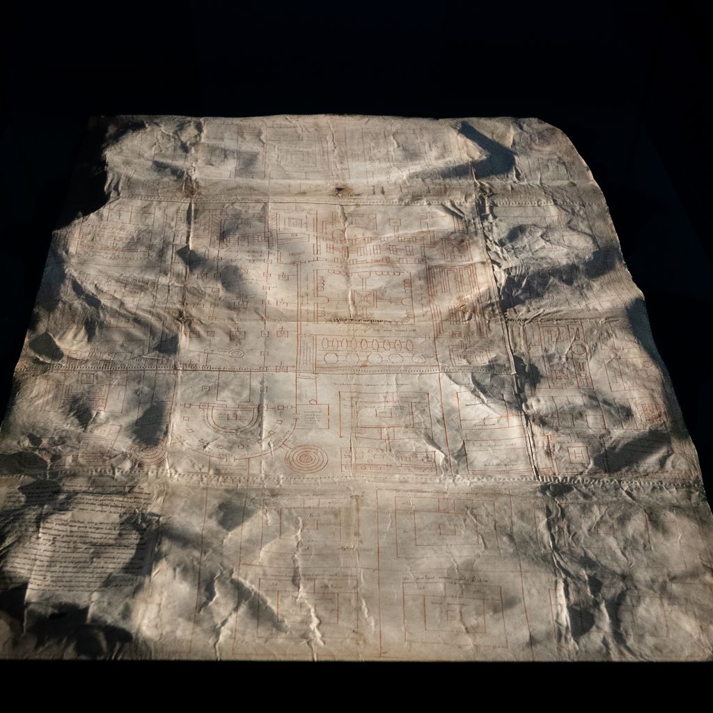 A large worn parchment covered with red architectural lines and inscriptions lies against a black display surround, with folds and losses visible.