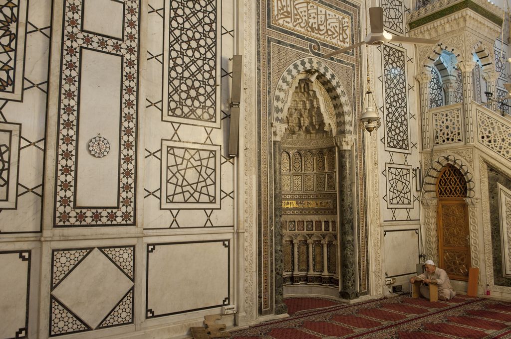 A richly framed mihrab niche stands in a geometric marble wall beside part of a stepped minbar, with carpet rows, lamps, fans and a seated visitor.