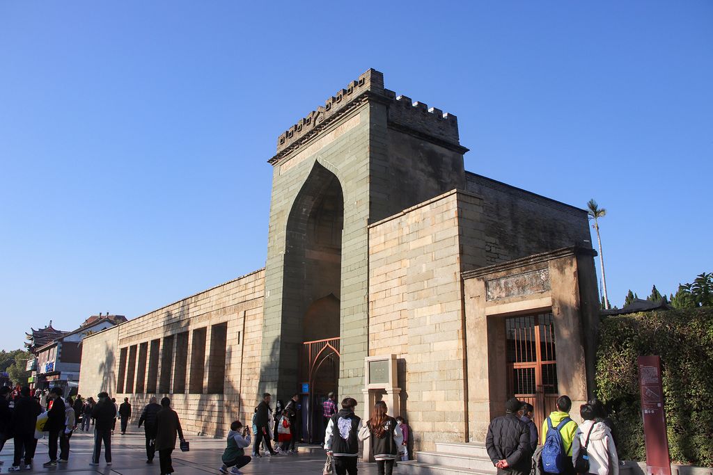 A high stone gateway with a pointed recessed arch stands beside a long wall and local tiled roofs on a busy paved street under blue sky.
