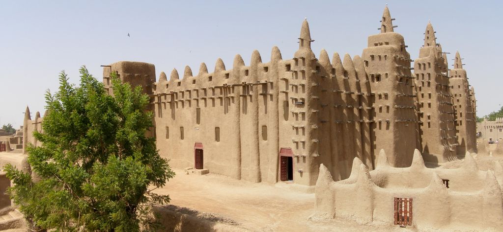 A monumental earthen mosque with three projecting towers, rows of timber poles and a raised enclosing wall stands in bright sun behind a leafy tree.