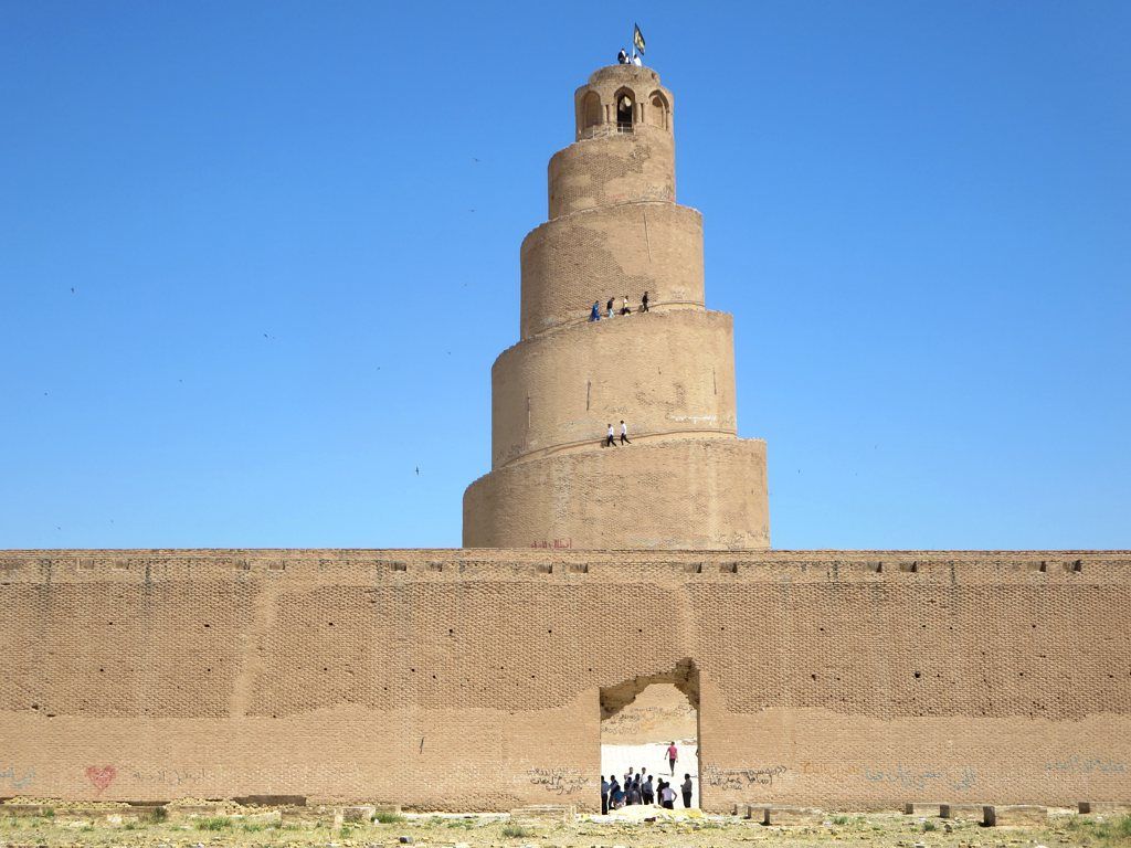 A broad spiralling brick tower rises behind a long mosque enclosure wall with a central doorway, with small visitors on the ramp and at the gate.