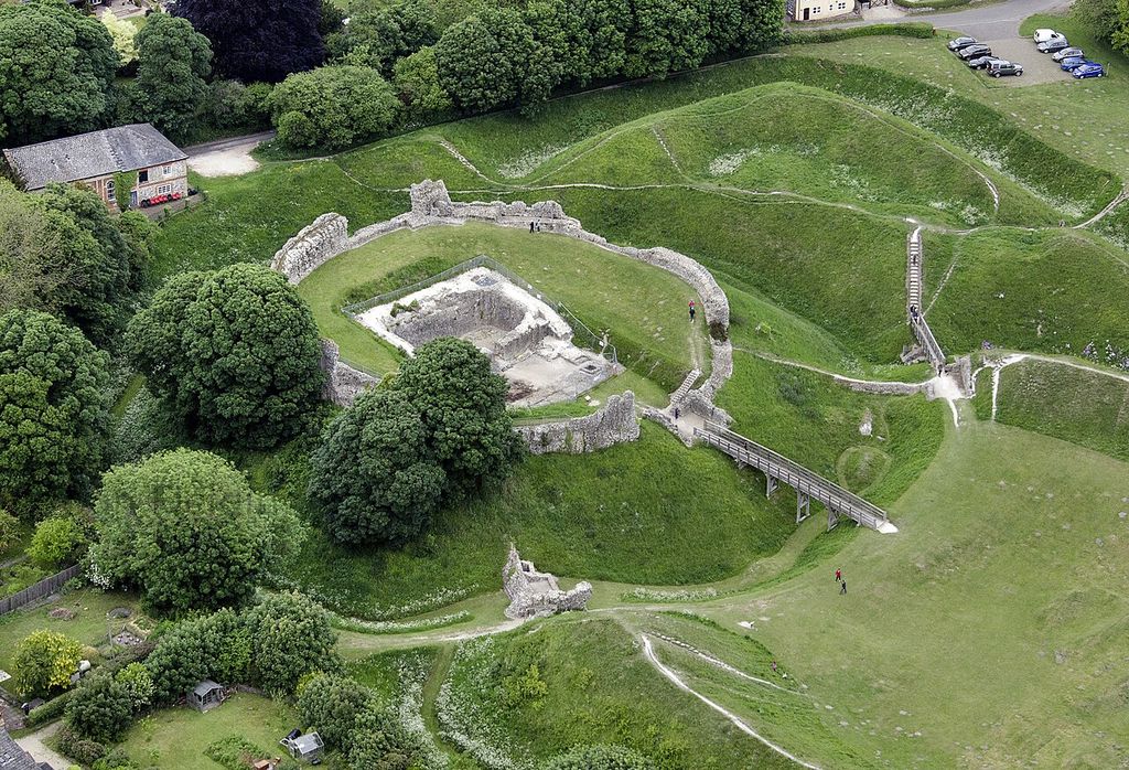 A raised walled enclosure containing ruined buildings is surrounded by deep ditches and grassed courts.