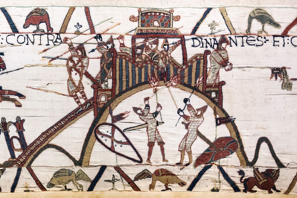 An embroidered tower and palisaded enclosure stand on a mound reached by a rising bridge, with armed figures around them.