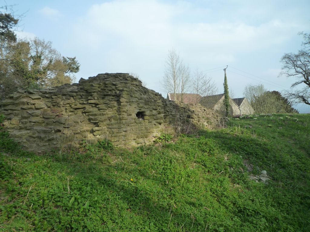 A low ruined stone wall crosses a grassy slope with part of a church behind trees nearby.