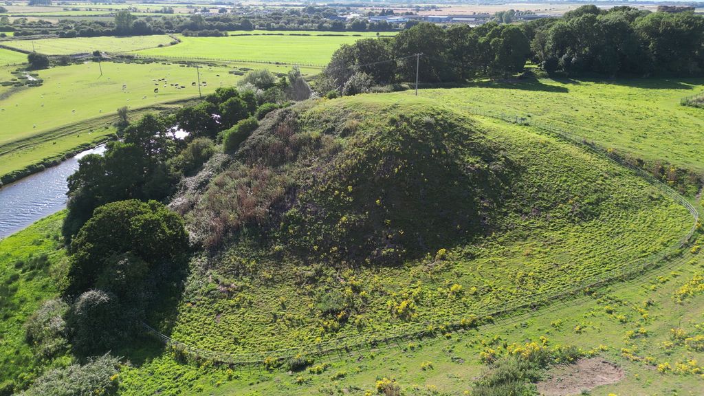 A broad grassy mound rises steeply beside a river within a fenced landscape.
