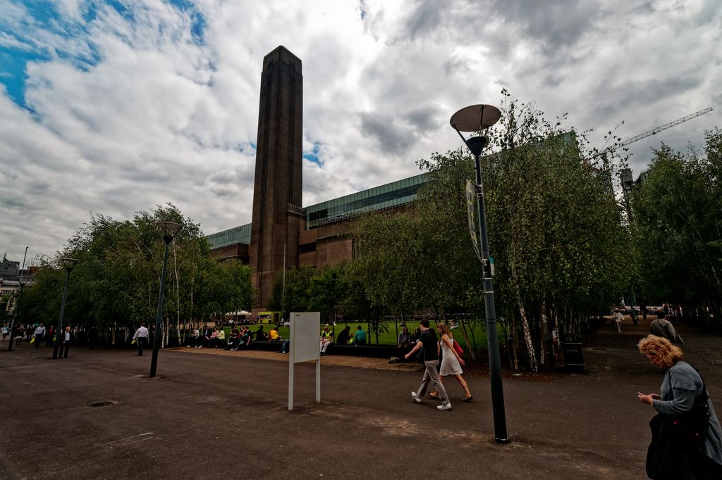 A tall square brick chimney and broad dark brick building rise behind trees and a public walkway, with a long glazed band at roof level.