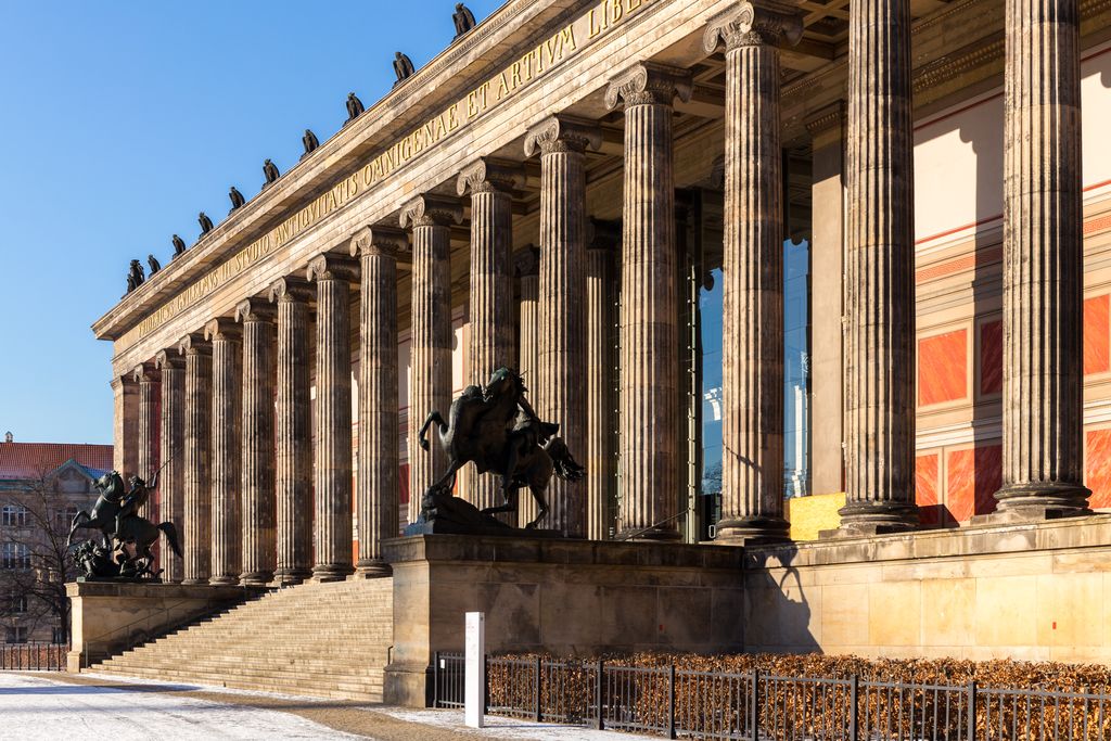 A long Ionic colonnade rises above a broad snow-edged stair in strong winter sunlight, with sculpture along the roof and monuments in front.