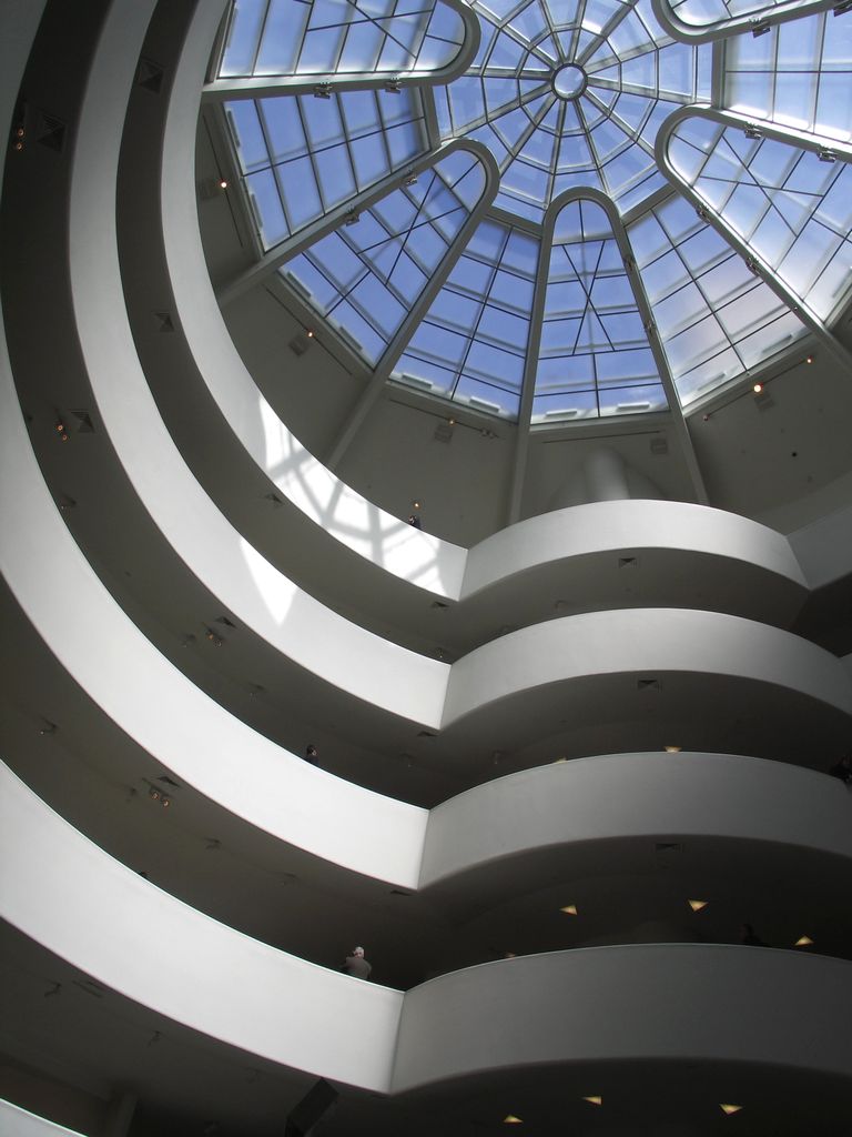 Smooth white ramps curve upward around a central void toward a radial glazed dome, with a few visitors visible on the lower levels.
