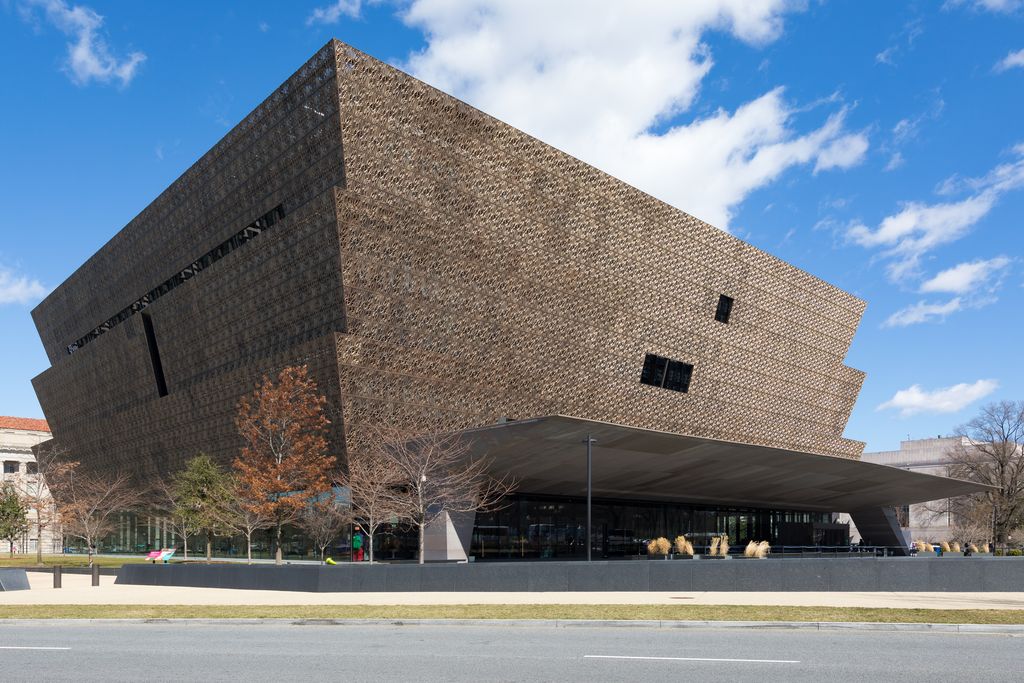 A three-tiered bronze-coloured museum with a dense patterned screen projects above a dark glazed base and broad entrance canopy under a blue sky.