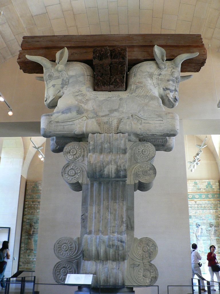 A colossal carved double-bull capital supports reconstructed dark timber beams in a museum gallery, with visitors and glazed brick panels behind.