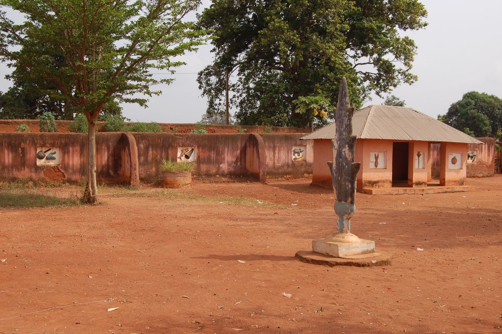 A broad red-earth court contains a low relief-decorated wall, small rendered building, trees and a tall freestanding sculptural form.
