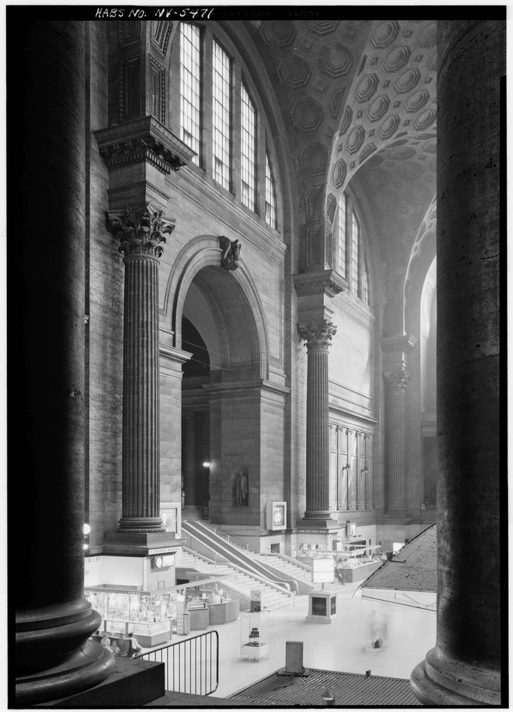 A black-and-white survey photograph looks between huge columns into a monumental station waiting room with high arched windows, stairs, kiosks and a coffered vault.