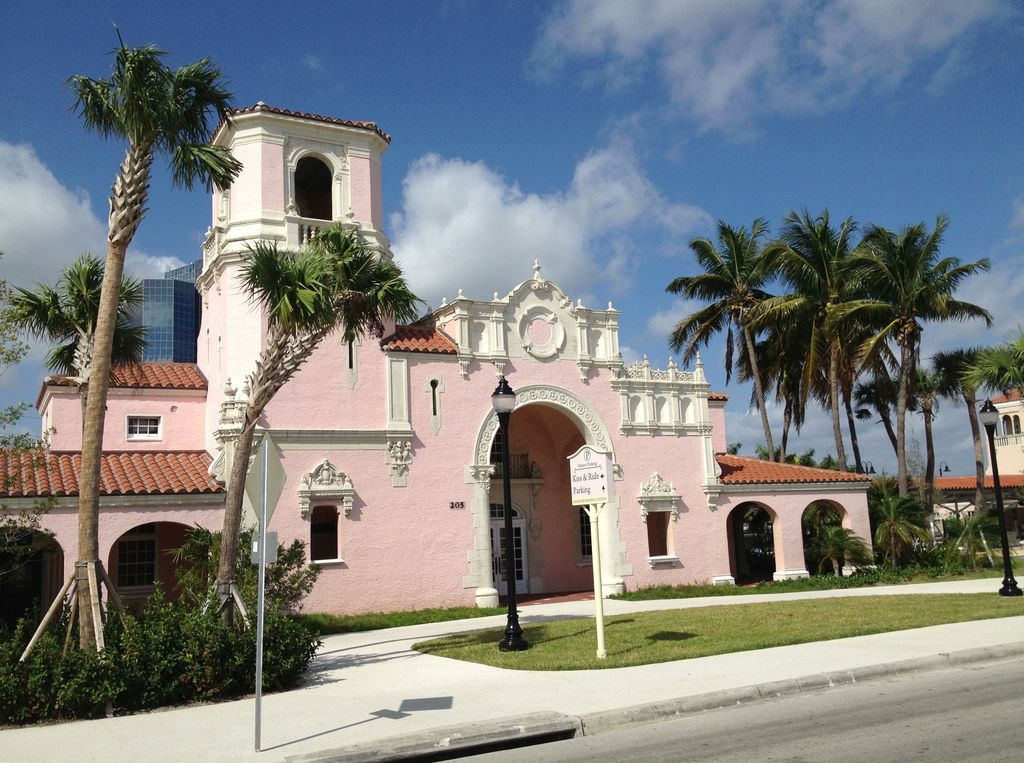 A pink stucco station with tiled roofs, a square entrance tower and open arcades stands among palms beside a street.