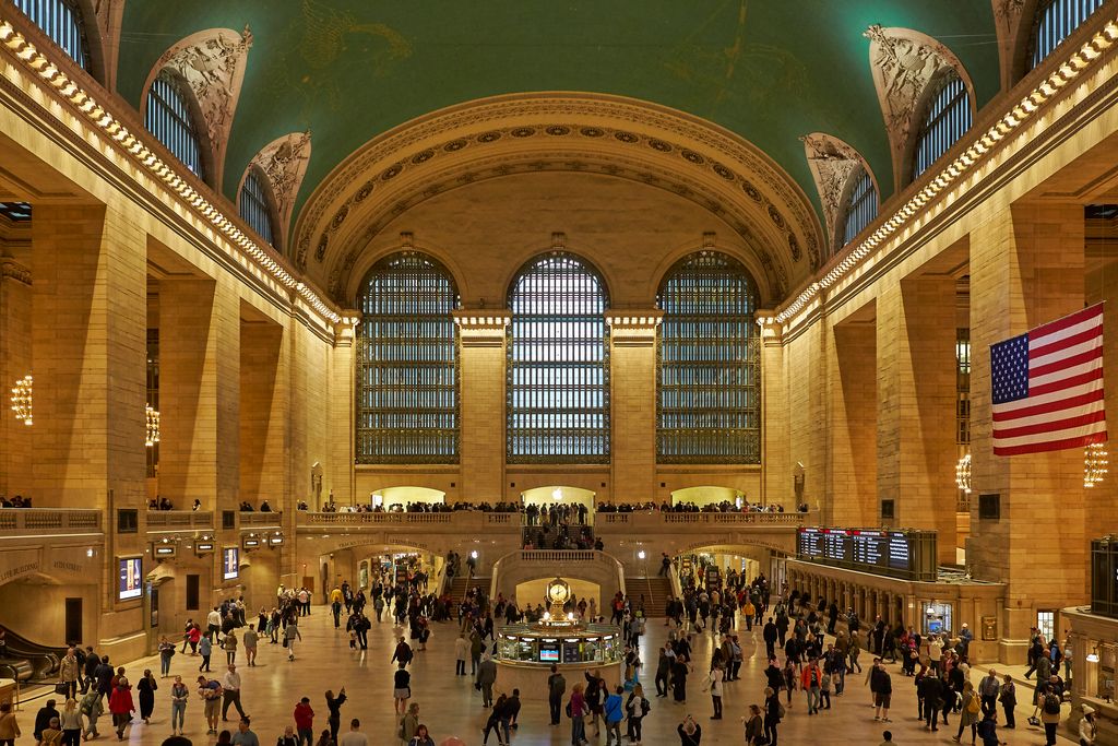 Crowds cross a high stone concourse beneath a turquoise vaulted ceiling, tall windows and chandeliers around a central information booth and clock.