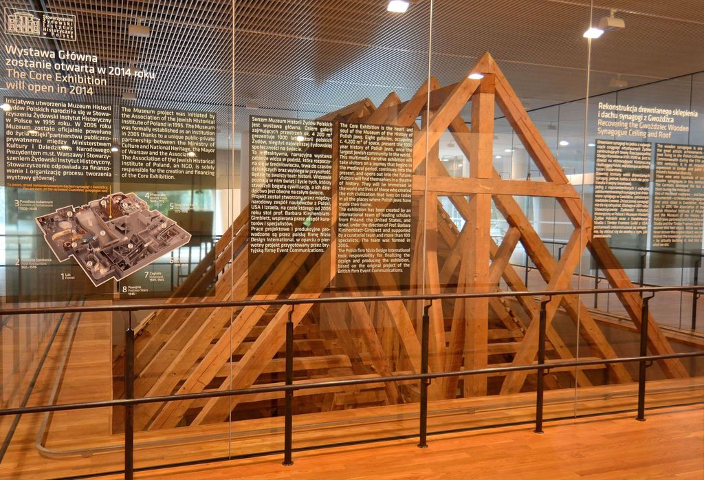 New timber trusses and braces stand behind museum glass and railings beside explanatory panels and a model of the exhibition.