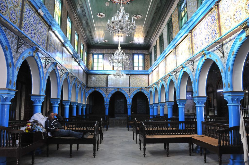 A long blue-arcaded hall with patterned tiles, high windows, chandeliers and benches forms a central passage around a seated visitor.