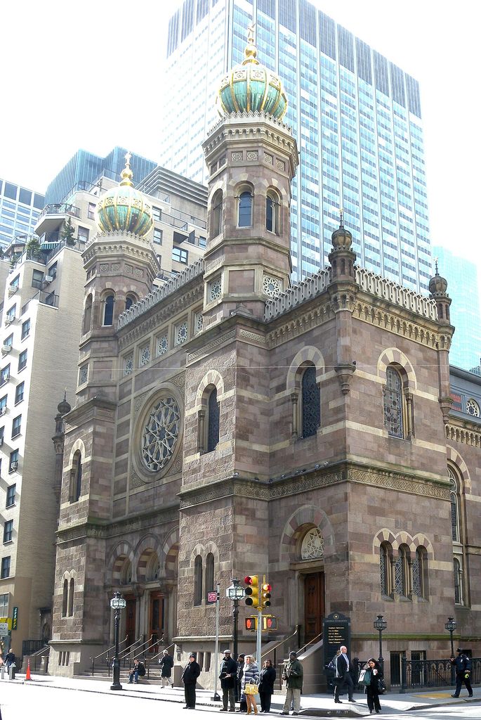 A banded masonry synagogue with twin onion-domed towers, a large circular window and arched doors stands amid tall Manhattan buildings and street traffic.
