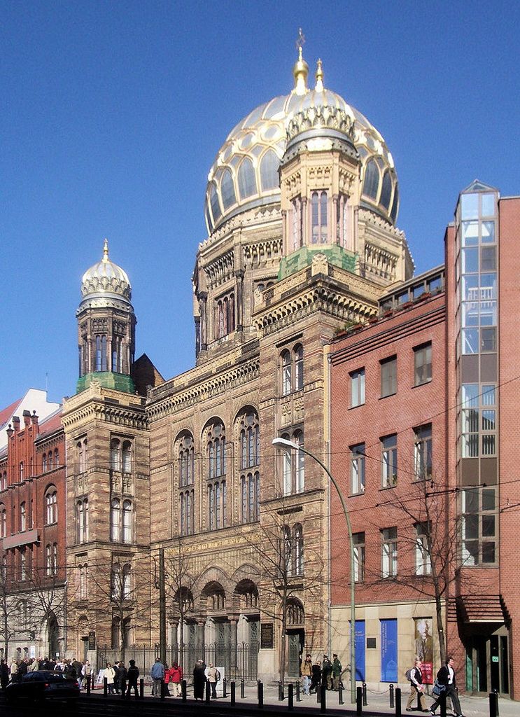 A richly patterned stone street façade with tall arched windows, two small domed towers and a large ribbed dome stands beside later brick buildings.