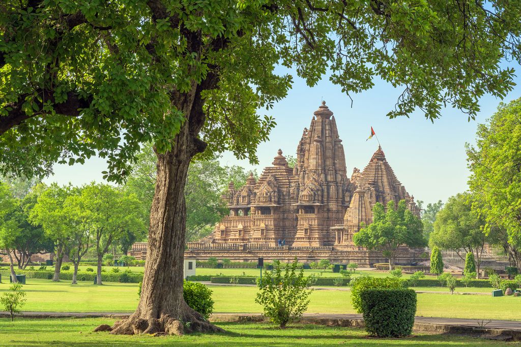 A tall clustered stone temple rises above a landscaped garden, with a principal shikhara and lower roof masses framed by a large tree.