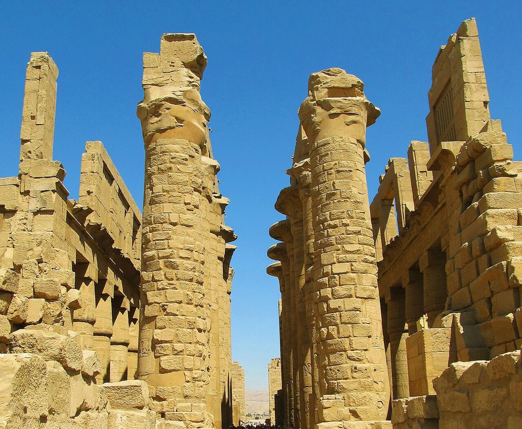 Massive carved sandstone columns and beams rise around tall clerestory openings against a deep blue sky, with large roof sections missing.