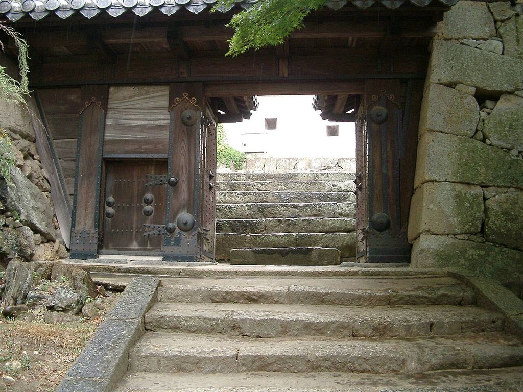 Heavy dark timber gate leaves with metal fittings open onto pale stone steps between rough masonry walls.