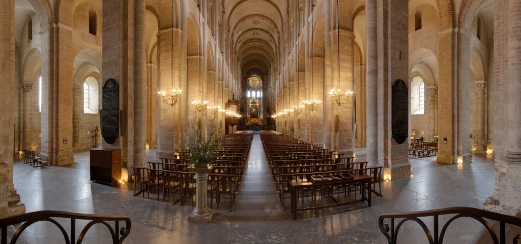 A long church nave has repeated round arches at two levels and across its high curved vault above rows of chairs.