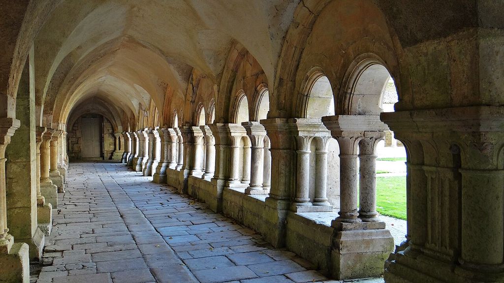 A stone-paved covered walk recedes beside paired columns and round arches opening toward a bright grass court under a vaulted roof.