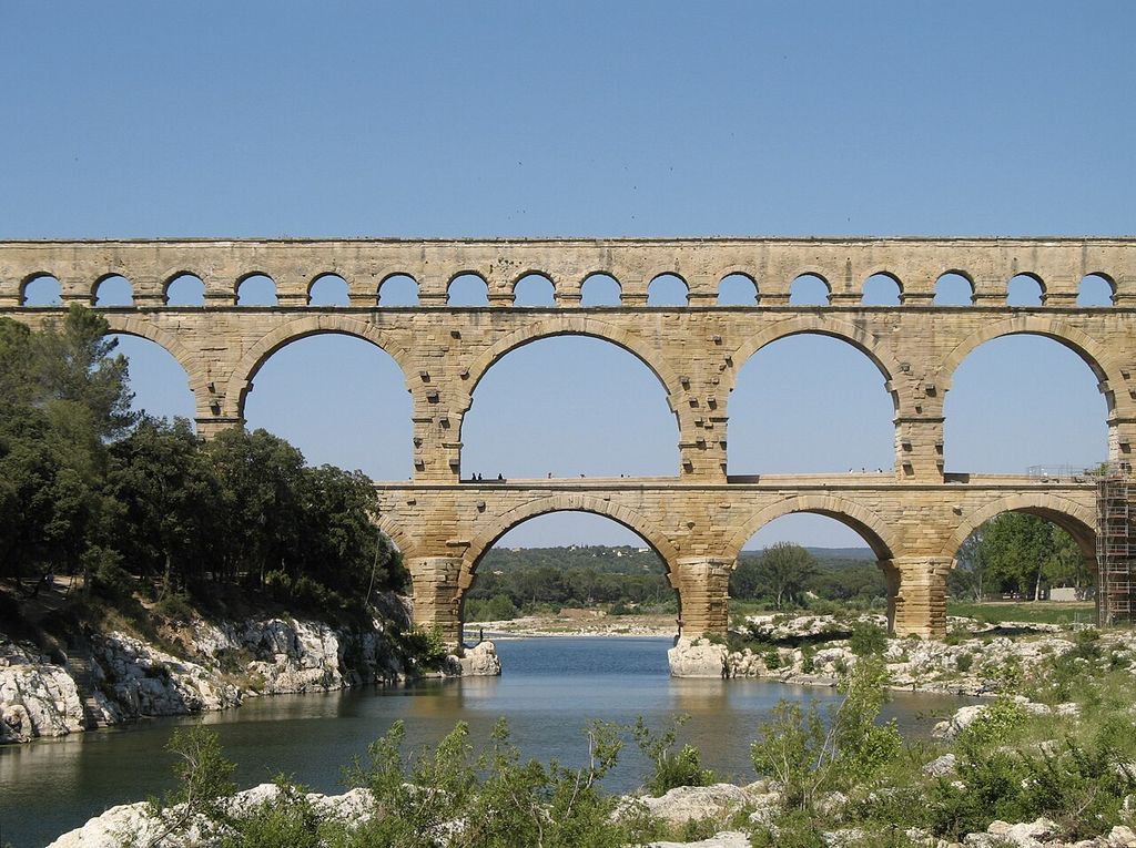 Three levels of pale stone arches cross a river valley, with broad lower spans, smaller upper bays, trees and scaffolding at the far right.