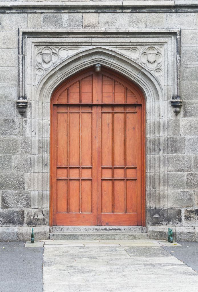 A broad pointed stone doorway with steeply curved shoulders surrounds wooden double doors beneath carved spandrels and a horizontal hood mould.