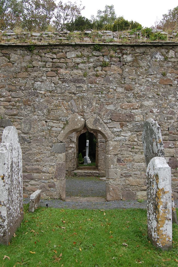 Two curved stones form a pointed ogee doorway beneath a separate shallow arch in the rubble wall; another doorway is visible beyond.