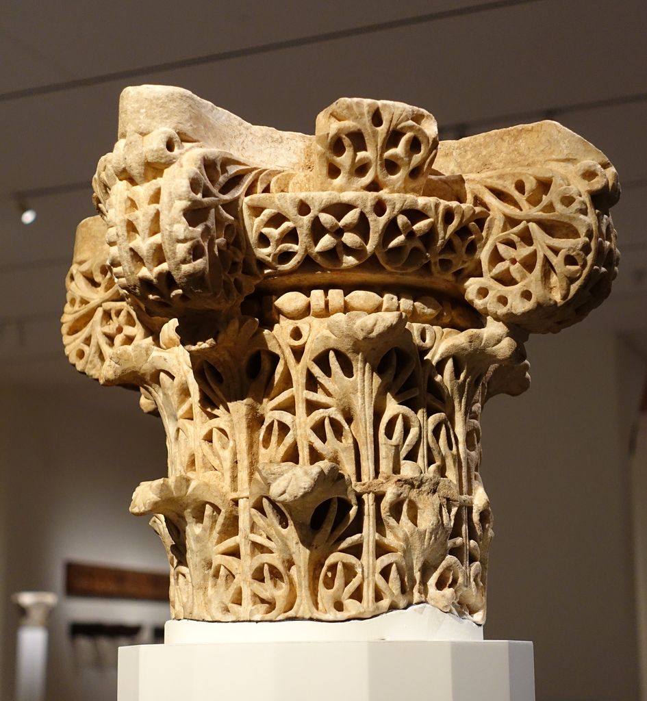 An intricately pierced marble capital has scrolling vegetal forms on four sides and a projecting curved abacus above.