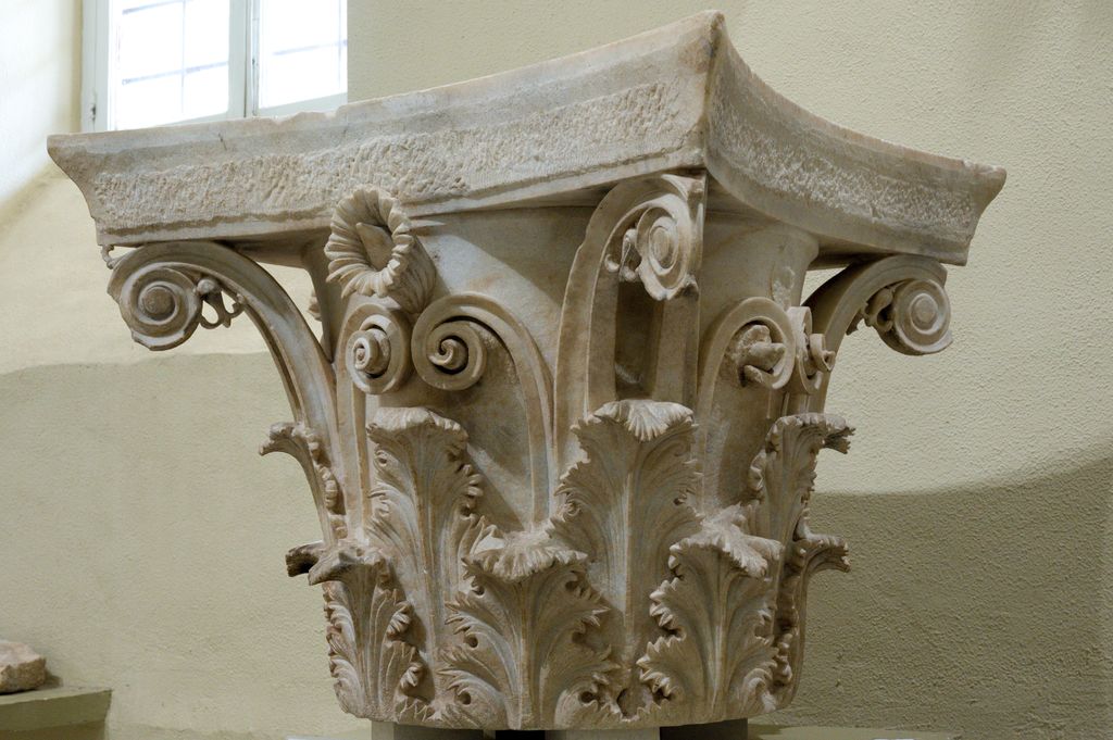 A marble capital has two tiers of deeply carved leaves, rising stalks, spiral corner forms and a curved square abacus.