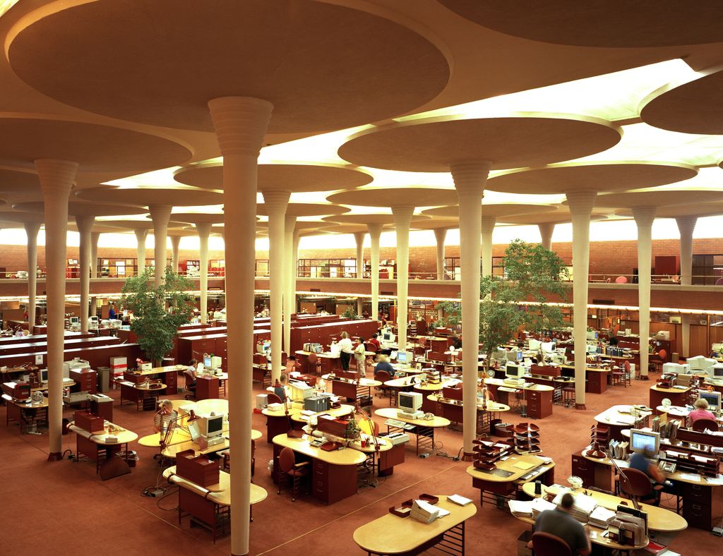 Slender columns widen into large circular ceiling pads above rows of occupied office desks.