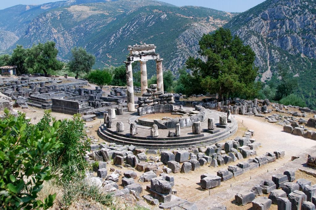 A circular ancient building with three reset columns stands among low ruins against steep mountains at Delphi.