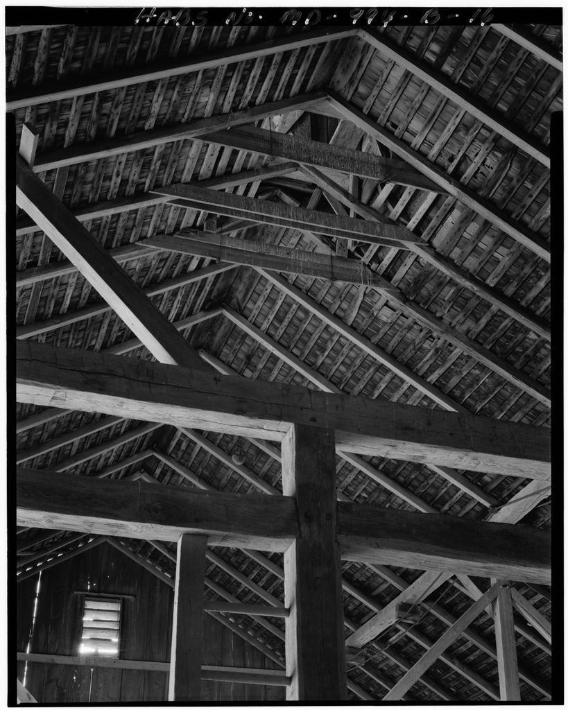 A monochrome interior view looks through heavy timber posts, beams, braces and roof trusses toward a framed opening beneath the cupola.