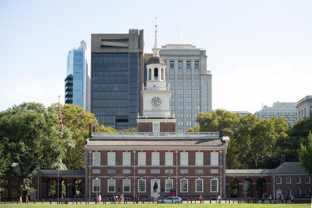 A symmetrical red-brick hall rises through a white clock stage, open cupola, small lantern, spire and weather vane against modern city towers.
