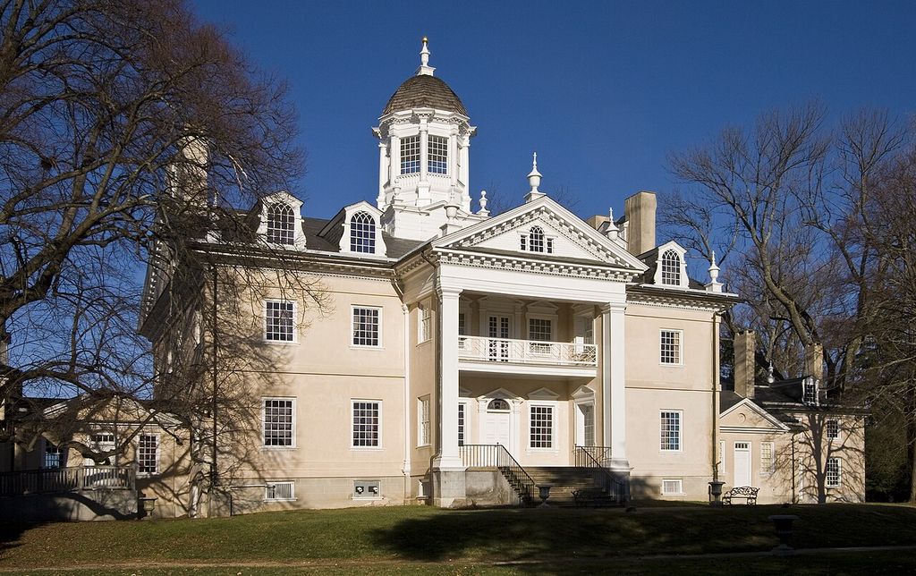 A pale Georgian mansion with a central glazed cupola, portico, dormers and wings stands on a winter lawn beneath a deep blue sky.
