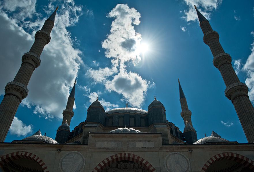 A lead-covered central dome and smaller roof volumes rise between four tall minarets against a bright blue, cloud-filled sky.