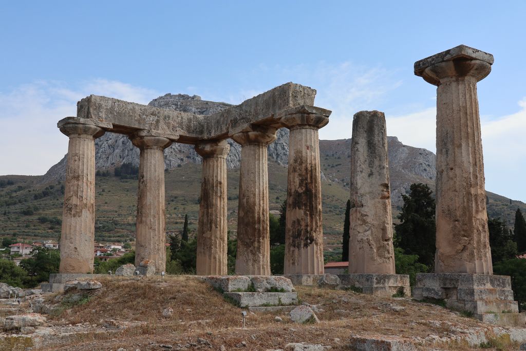 Seven weathered fluted columns with broad spreading capitals stand on a raised platform against Acrocorinth.