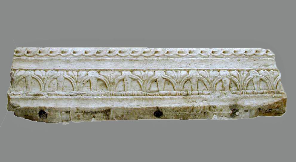 A long detached marble block has a projecting upper edge, repeated leaf-like carving, weathered mouldings and broken ends against grey.
