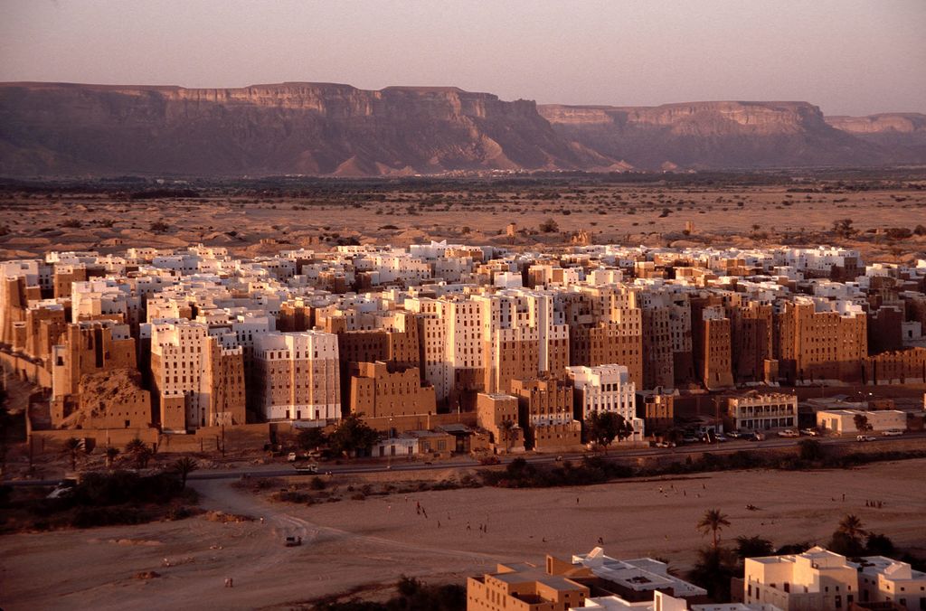 A dense cluster of tall brown and white earthen tower houses rises from a desert settlement beneath a broad plateau.
