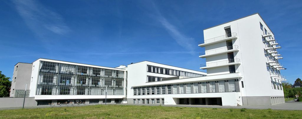 A long white modern complex combines a broad glass-grid workshop wing, a raised connecting bridge and a taller balcony block beside a lawn.